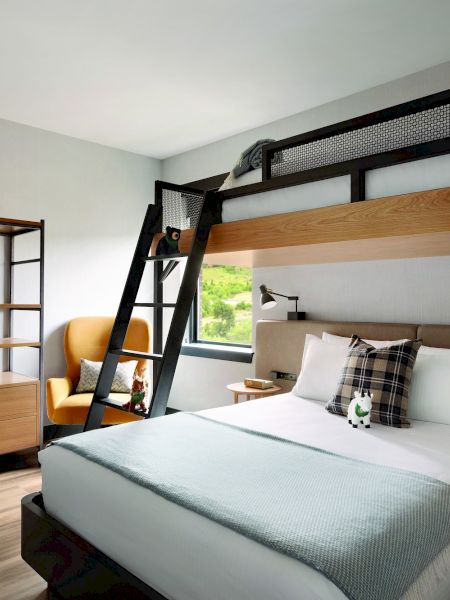 A modern hotel room with a double bed, a loft bed above it, a ladder, a seating area, a wooden closet, and bathrobes hanging.