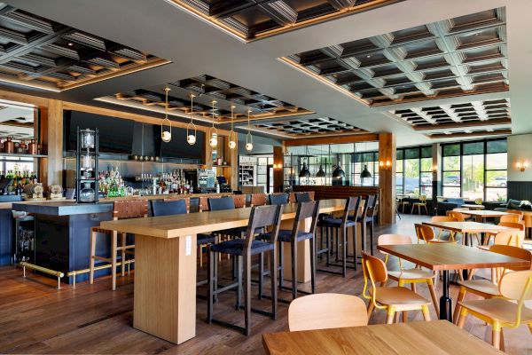 A modern restaurant interior featuring a bar with high chairs, wooden tables with chairs, stylish ceiling panels, and large windows letting in natural light.