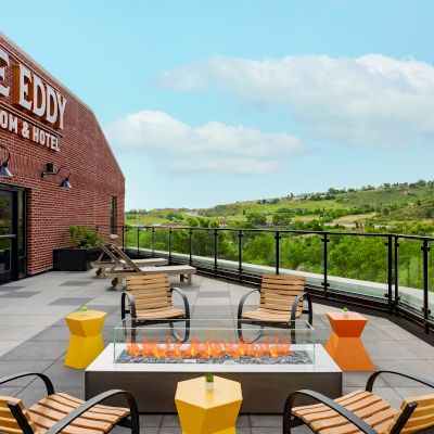 An outdoor patio at "The Eddy Taproom & Hotel" with seating around a fire pit, overlooking a scenic landscape with hills and greenery, under a blue sky.