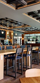 This image shows a modern, well-lit restaurant interior with wooden furniture, a bar area, and stylish ceiling design. Multiple seating options are available.