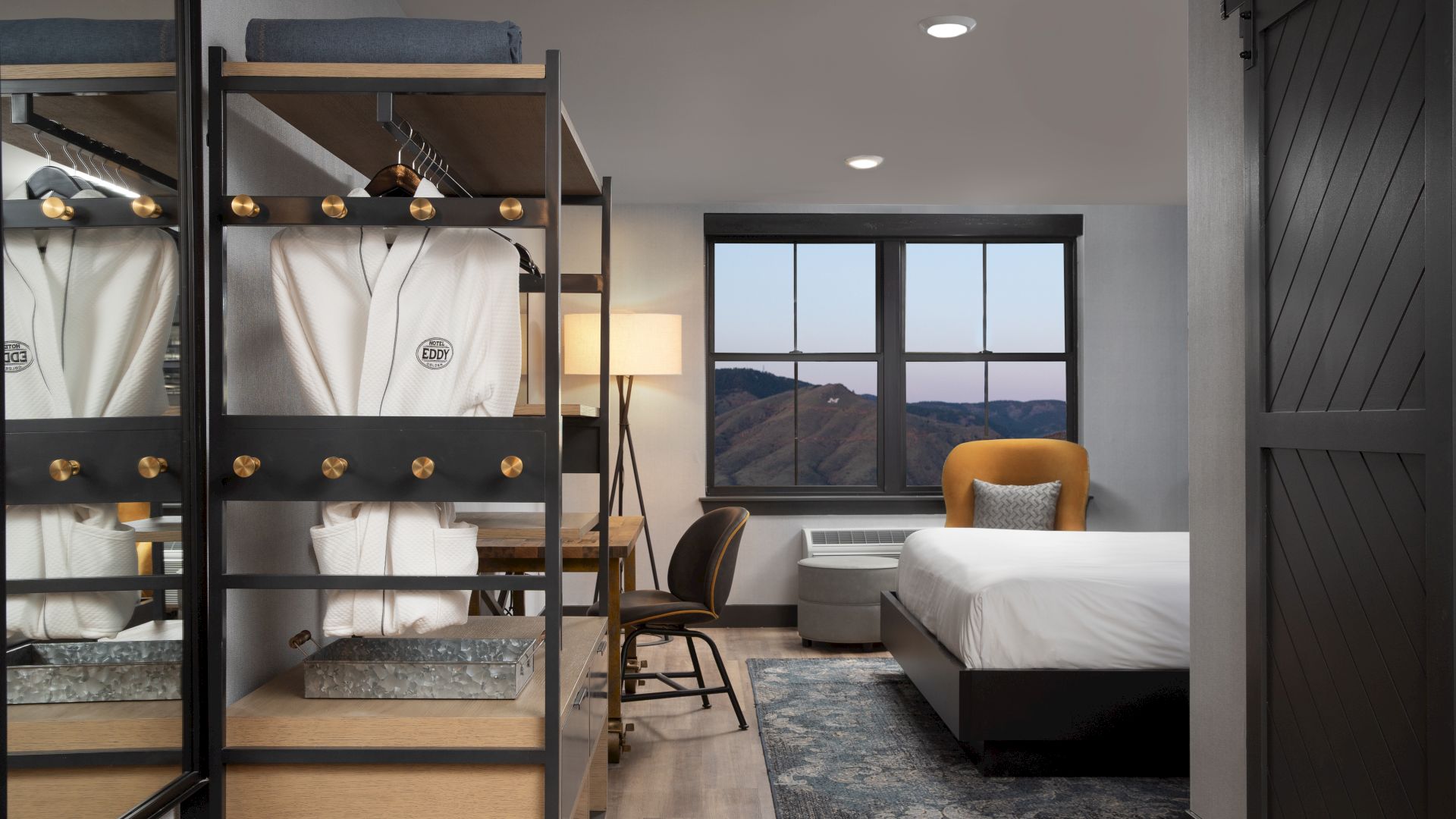 This image shows a modern hotel room with a bed, a desk with a chair, a wardrobe with robes, and a window with a mountain view at dusk.