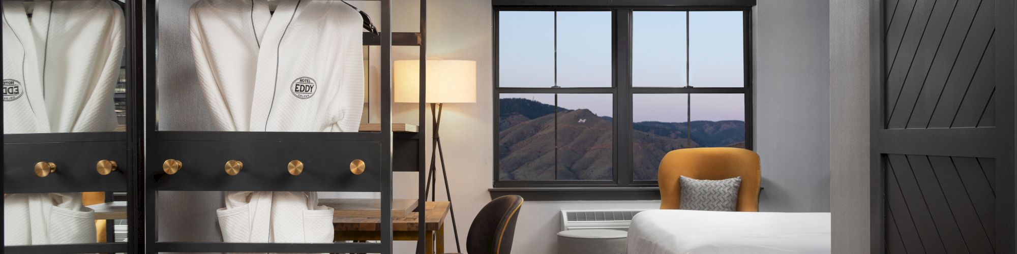 This image shows a modern hotel room with a bed, a desk with a chair, a wardrobe with robes, and a window with a mountain view at dusk.
