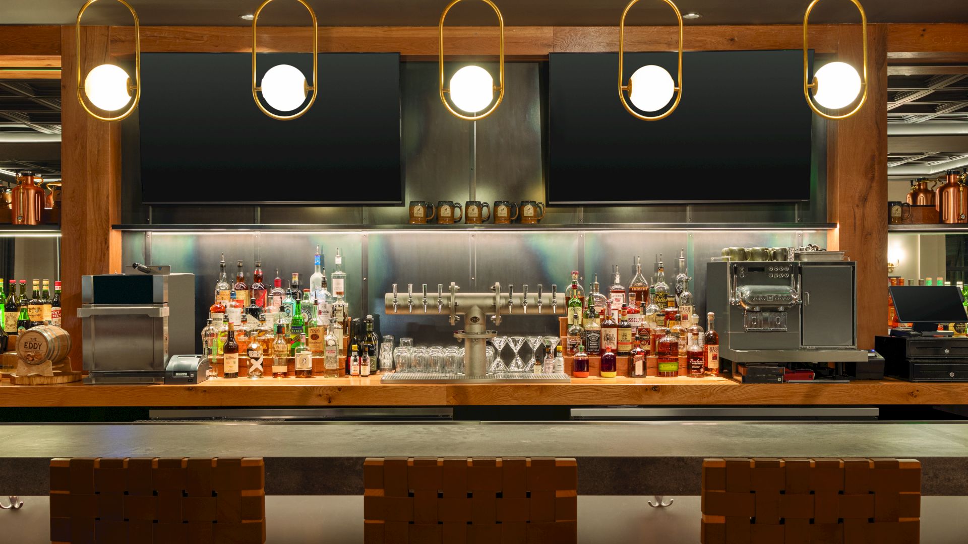 A stylish bar with a well-lit counter, liquor bottles, glassware, and bar stools ready for customers, plus a coffee machine on the right.