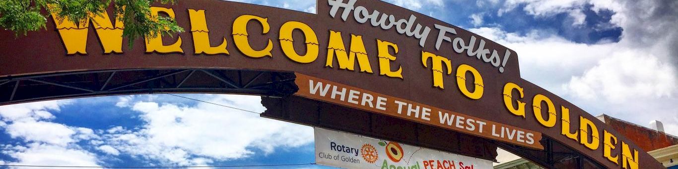 Welcome to Golden, a rustic sign arch reads &ldquo;Howdy Folks! WELCOME TO GOLDEN&rdquo; against blue sky, with a banner below about a Peach Sale.