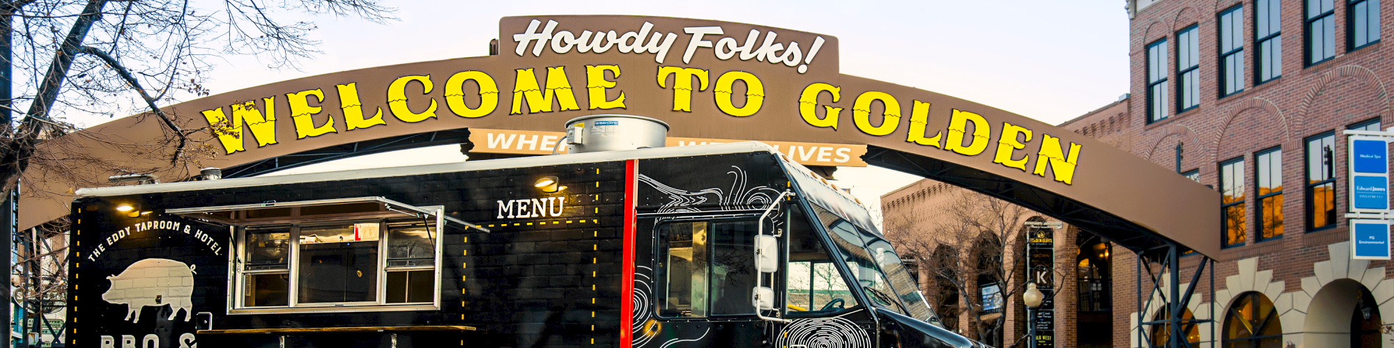 A food truck is parked under a "Welcome to Golden" archway on a street with buildings and a leafless tree in the background.