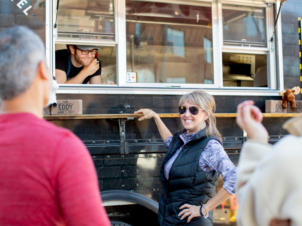 People line up for food from a black food truck; a smiling woman in sunglasses chats with the driver while others wait, ready to order.