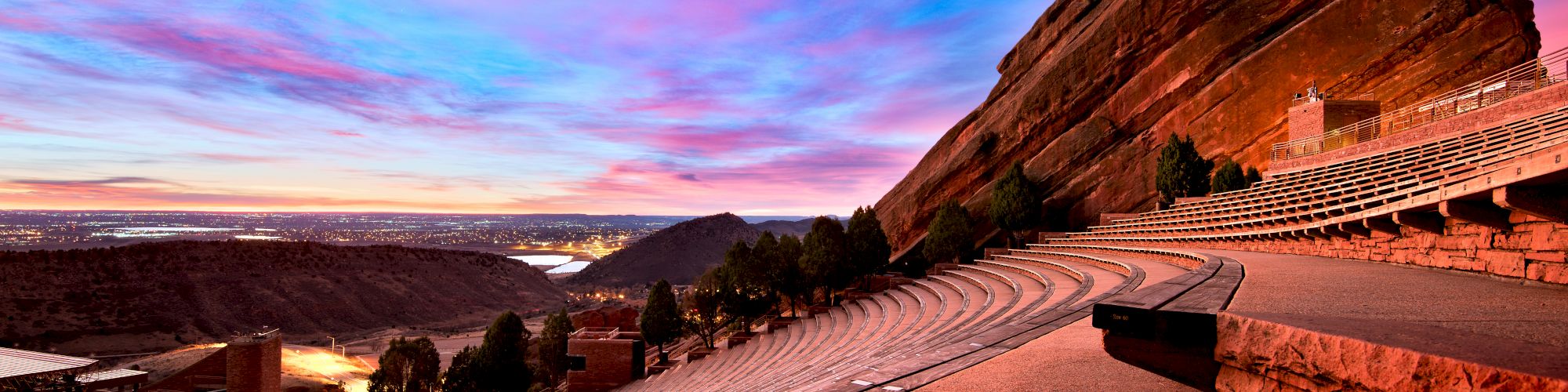 The image shows an outdoor amphitheater with large rock formations and a colorful sunset sky in the background.