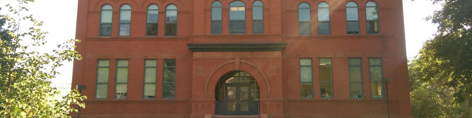 The image shows a red brick building with arched windows and a central entryway, surrounded by green lawns and trees with bicycles outside.
