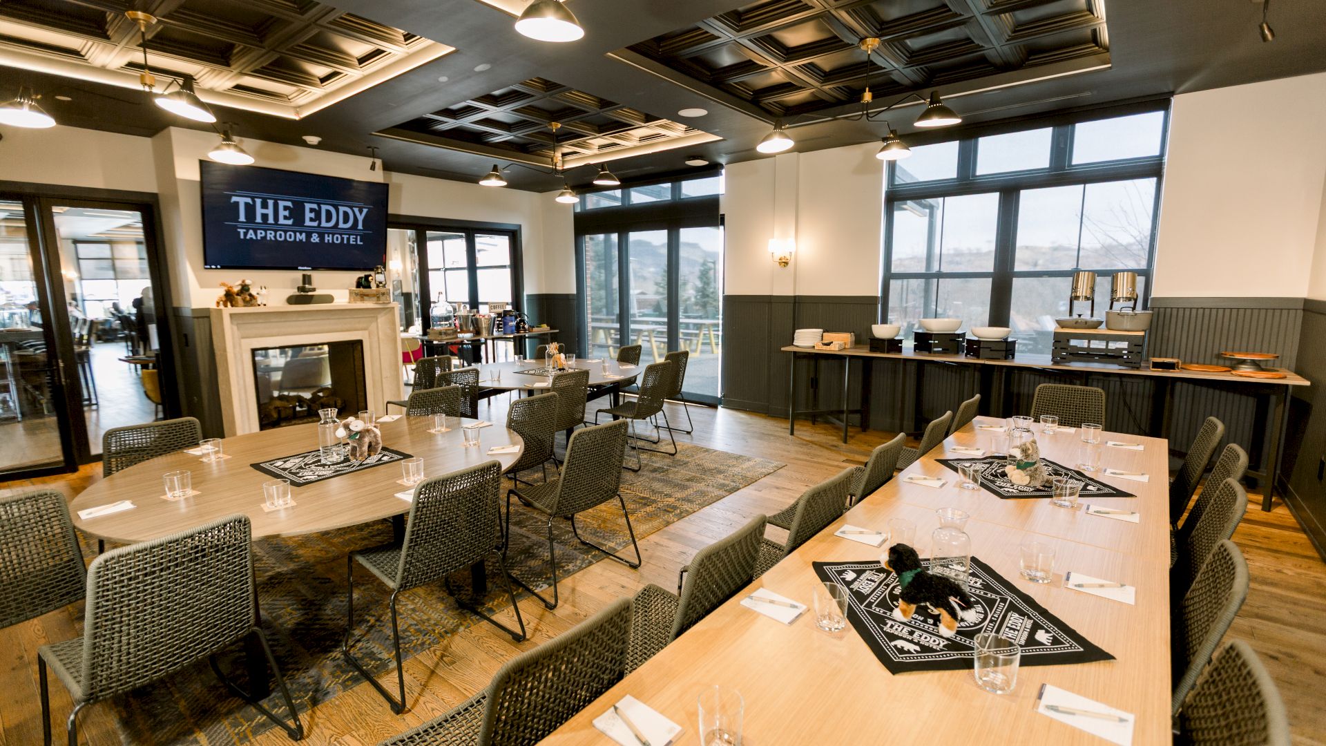 A modern event space with long wooden tables, black chairs, a stage area with a sign saying &ldquo;The Eddy,&rdquo; a fireplace, and large windows letting in daylight.