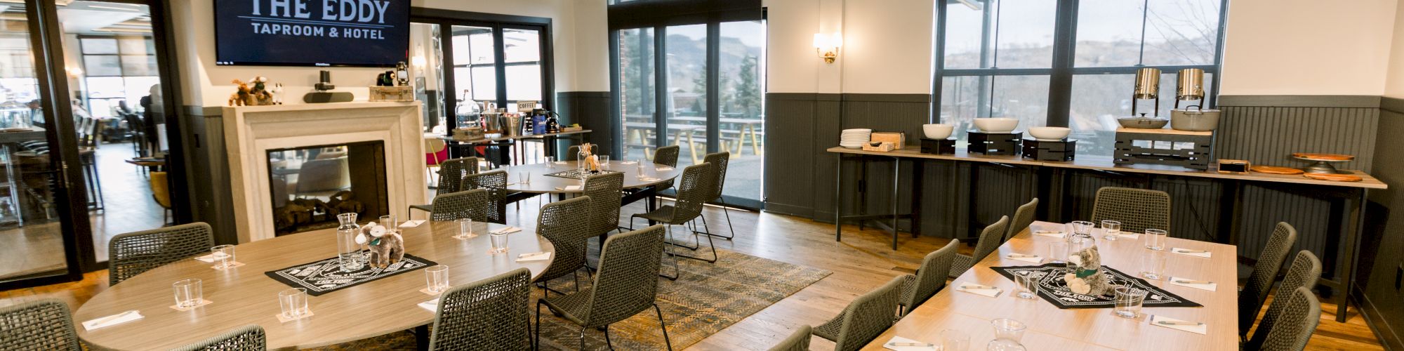 A modern event space with long wooden tables, black chairs, a stage area with a sign saying &ldquo;The Eddy,&rdquo; a fireplace, and large windows letting in daylight.