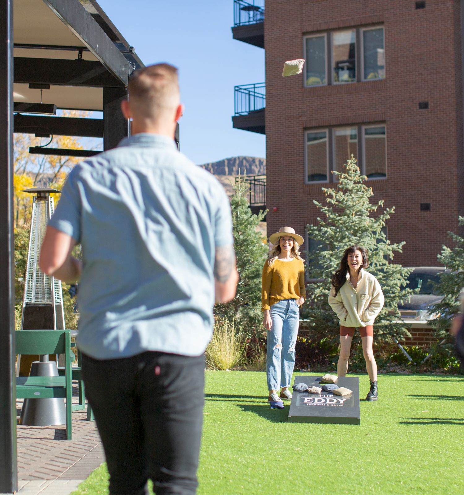 People playing cornhole on a sunny outdoor terrace near a brick building, with others watching and a man in a beige jacket preparing to throw.