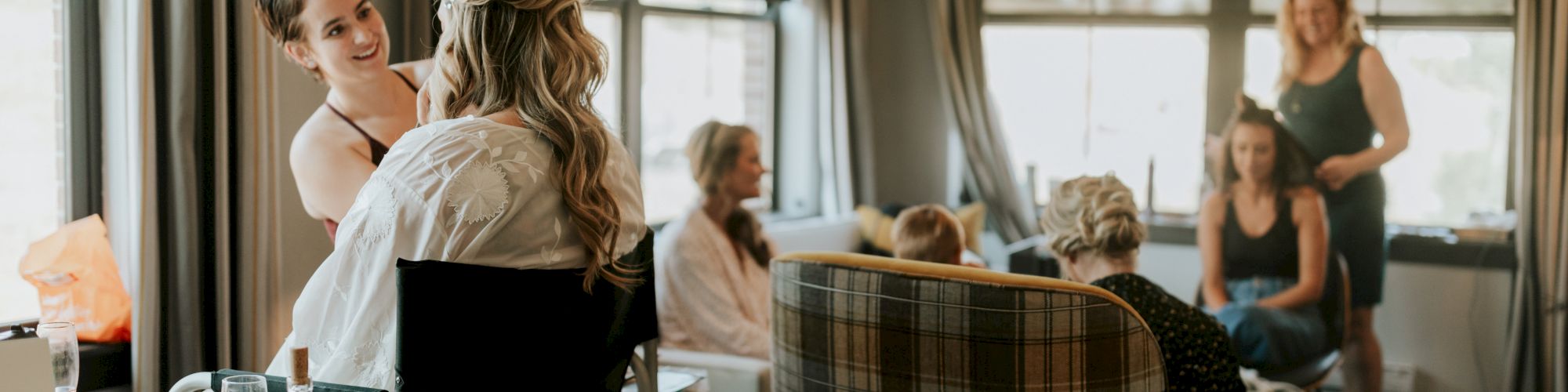 A stylist is doing makeup for a woman in a chair, while other women socializing in a cozy room with large windows and ample daylight.