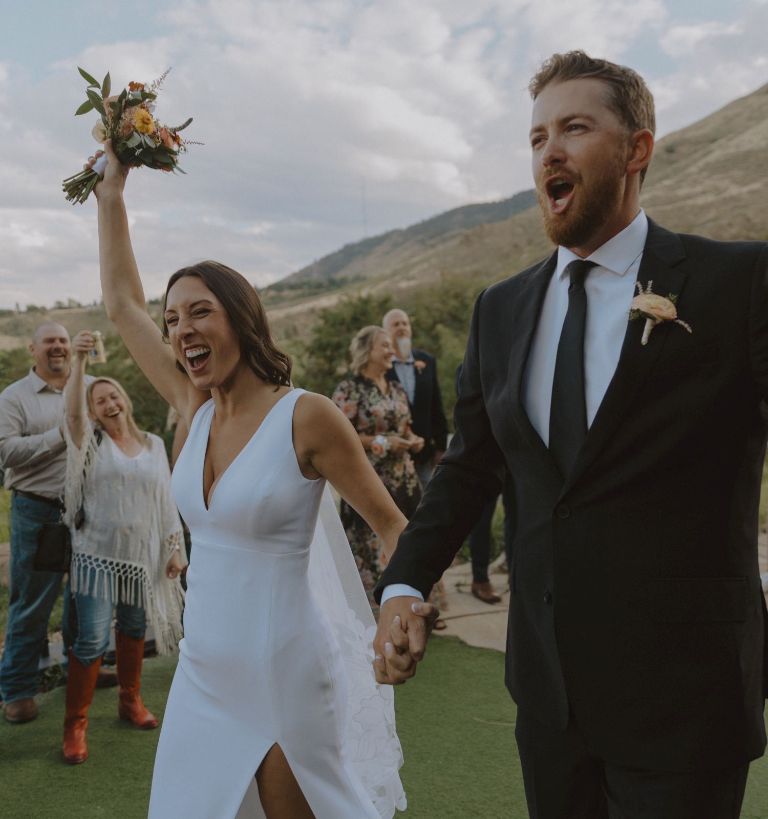 A bride cheers with a bouquet while holding hands with her groom, both celebrating outdoors in front of mountains in Golden Colorado at The Eddy Taproom & Hotel