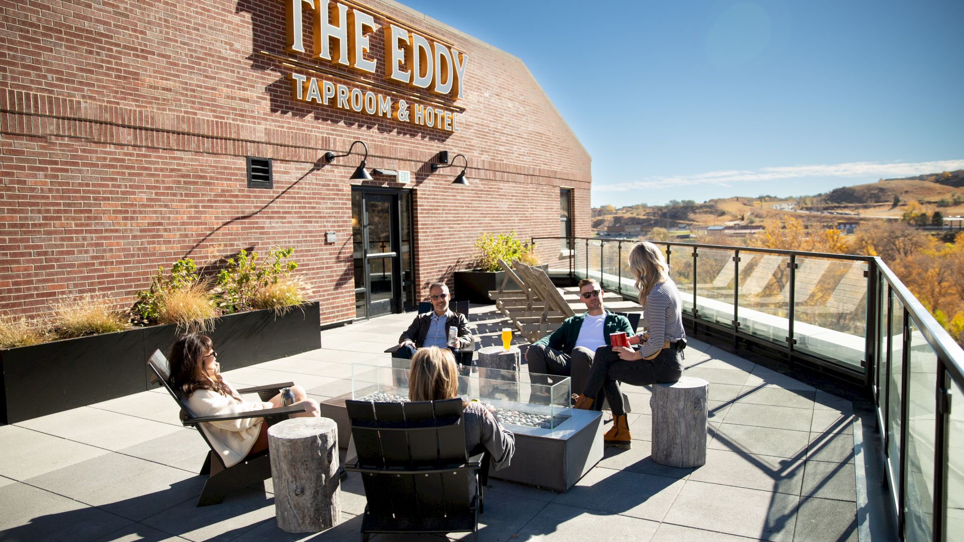 A group of people relaxing on a rooftop patio at "The Eddy Taproom & Hotel" with a scenic view of the landscape in the background.