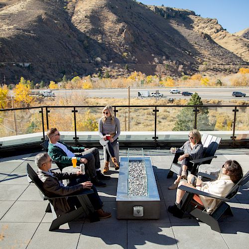 A group of people sits around an outdoor fire pit on a patio with mountains in the background, having a discussion and enjoying the sunny weather.
