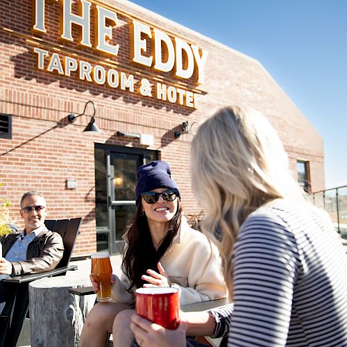 Three people are outdoors at "The Eddy Taproom & Hotel." They are enjoying drinks and conversing on a sunny day, with a brick building in the background.