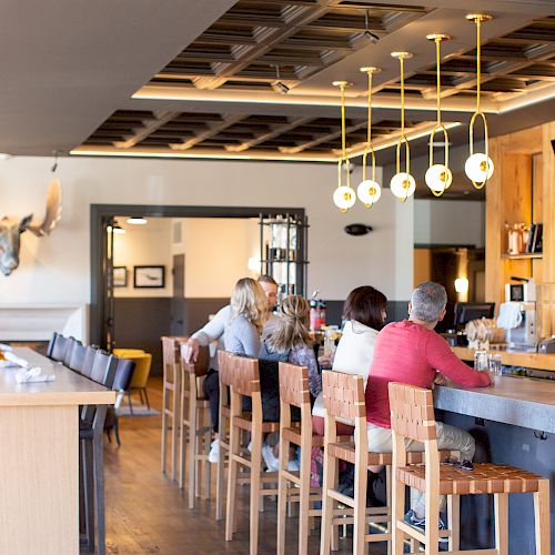 A group of people sitting at a stylish bar with wooden chairs and modern lighting, enjoying their time in a cozy, well-lit atmosphere.