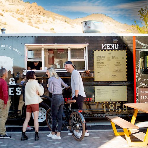 A group of people standing outside a food truck with menus on its side, ordering food; a picnic table is in the foreground.