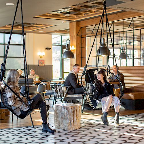 The image shows a modern café with hanging chairs, people sitting and chatting, wooden decor elements, and a cozy atmosphere.