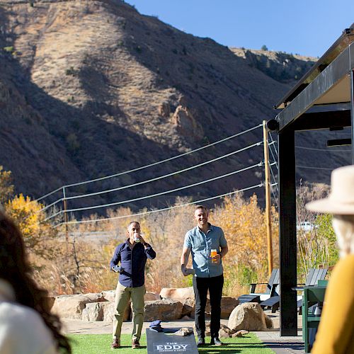 People playing a game outside with cornhole boards, set against a scenic mountain backdrop. Others watch, enjoying the sunny day.