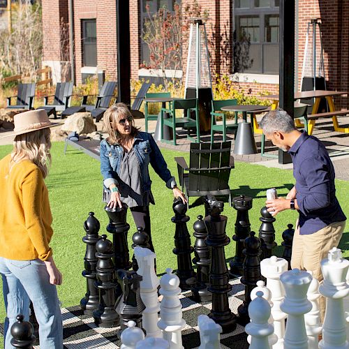 Three people are playing an outdoor game of chess with oversized pieces on a large chessboard near a building with a brick exterior.