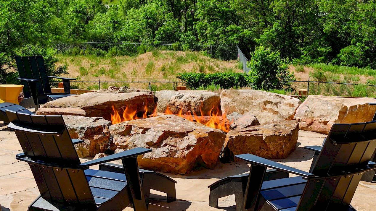 An outdoor seating area features black chairs arranged around a stone fire pit with a lush green backdrop, creating a cozy and inviting space.
