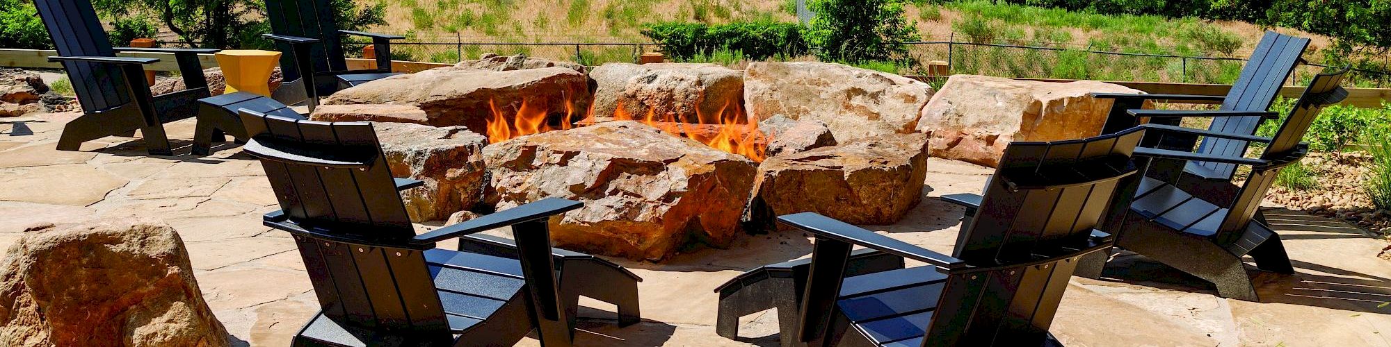 An outdoor seating area features black chairs arranged around a stone fire pit with a lush green backdrop, creating a cozy and inviting space.