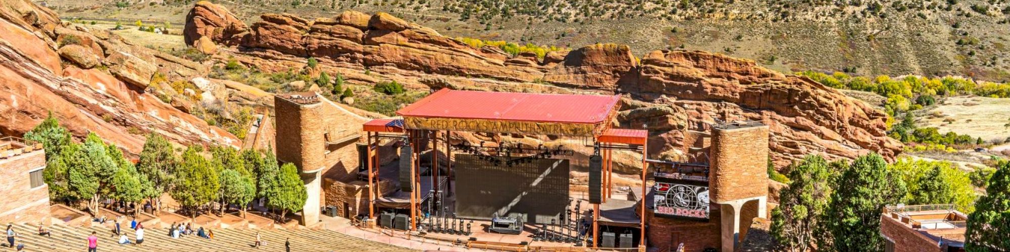 An outdoor amphitheater set within red rock formations, featuring a large stage area, with people walking and sitting in the seating area.
