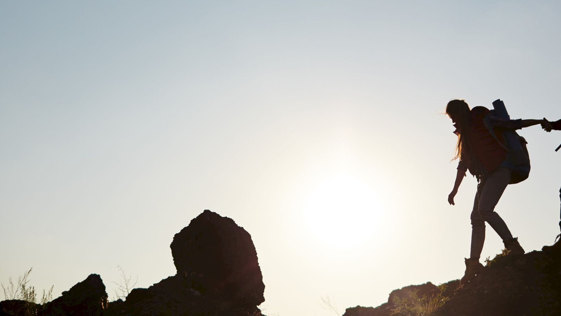 Silhouette of two hikers with backpacks trekking uphill against a sunset background, one helping the other climb.