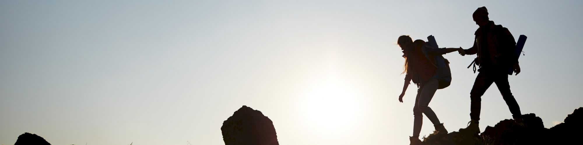 Silhouette of two hikers with backpacks trekking uphill against a sunset background, one helping the other climb.