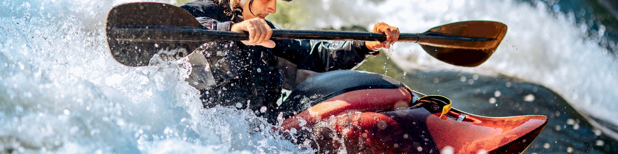 A person is seen kayaking through white water rapids, wearing safety gear including a helmet, while paddling vigorously to navigate the rough waters.