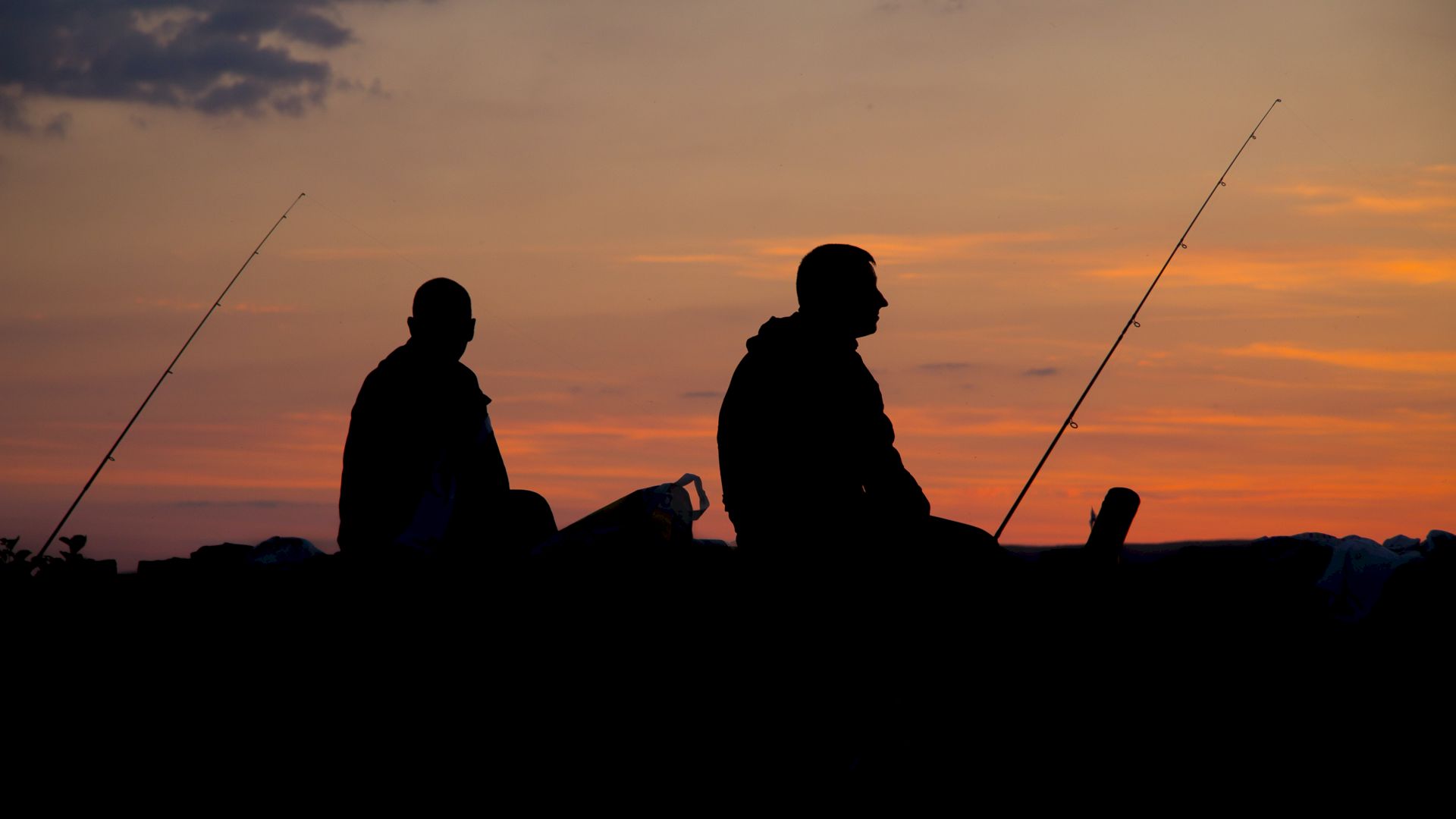 Two people are silhouetted against a sunset sky while fishing, with their fishing rods resting beside them.