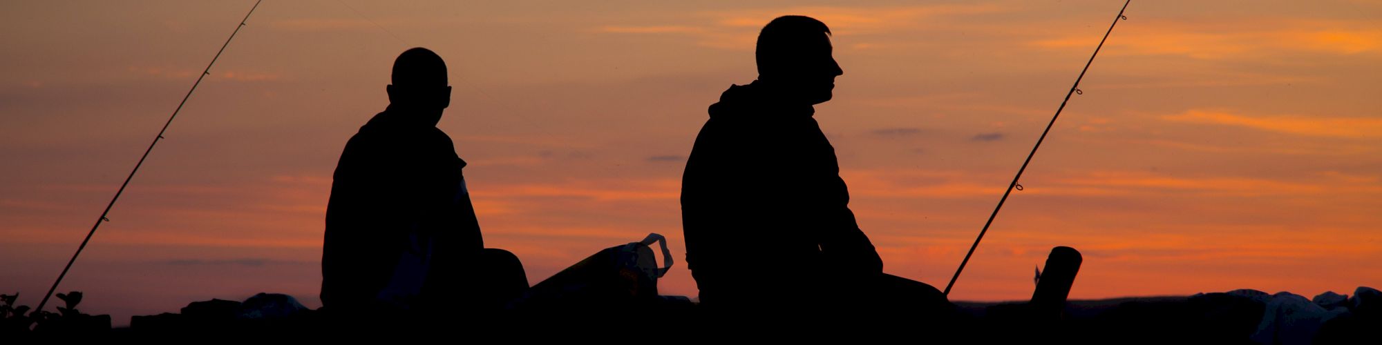 Two people are silhouetted against a sunset sky while fishing, with their fishing rods resting beside them.