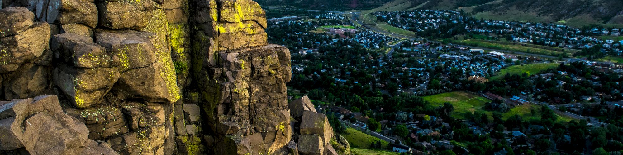 A scenic view of a rocky cliff with a lush green valley and small town below, set against a backdrop of rolling hills and a colorful sunset sky.