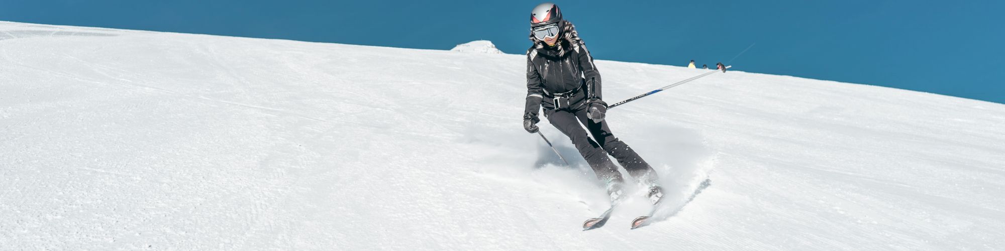 A person wearing ski gear glides down a snowy slope against a clear blue sky background.