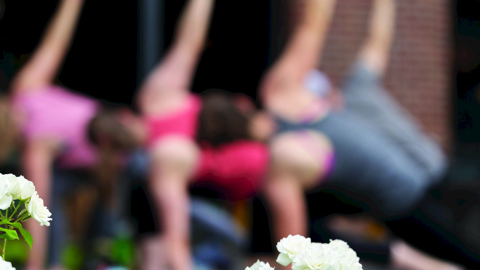 The image shows multiple people in a yoga pose, blurred in the background, with white flowers in focus in the foreground.