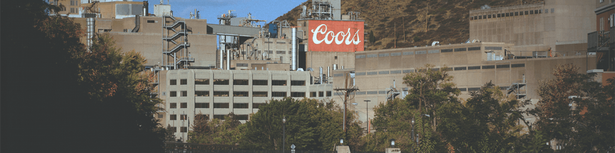 The image shows a large industrial facility with a sign labeled "Coors" in red letters, situated near a hilly landscape and a bridge over a river.