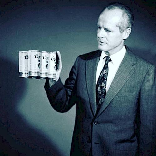 A man in a suit holds a pack of Coors Light beer cans, looking at them with a focused expression, in a monochromatic image.