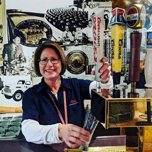 A person stands behind a bar, smiling and pouring a beer from a Coors Light tap. The background features vintage beer-related advertisements and memorabilia.