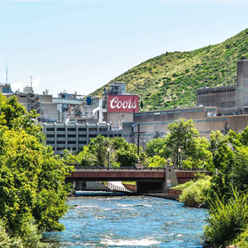 A river flows under a bridge with the Coors brewery in the background, surrounded by lush greenery and hills under a clear sky.