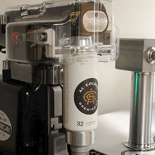 The image shows a can seaming machine with a can of AC Golden Brewing Company beer, next to a beer tap, likely used for brewing or canning beer.