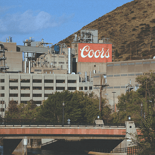 This image shows an industrial building complex with a prominent "Coors" sign, situated near a bridge and surrounded by trees and hills in the background.