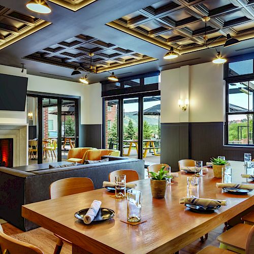 A modern restaurant interior with a wooden dining table, neatly set for guests, a cozy fireplace, large windows, and stylish lighting fixtures.
