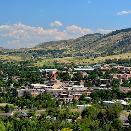 A scenic town nestled among green hills and mountains under a blue sky with clouds, featuring a mix of buildings and lush greenery.