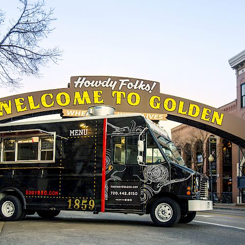 A food truck serving BBQ & shakes is parked in front of a "Welcome to Golden" sign in a city street scene, with buildings and a tree nearby.