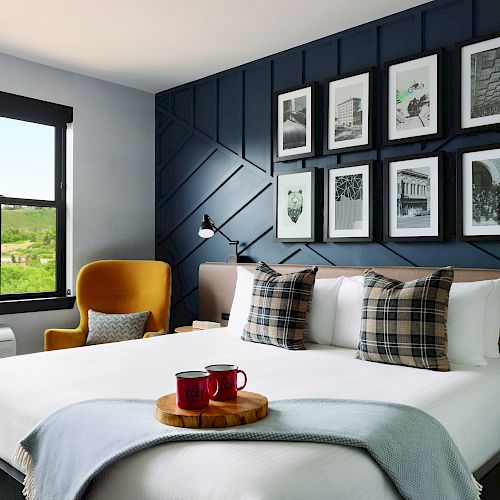 A cozy bedroom featuring a large bed with patterned pillows, framed artwork on the wall, a seating area with a yellow chair, and a scenic view outside the window.