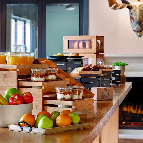 This image shows a buffet with fruits, juices, pastries, and desserts arranged on a wooden counter, set in a cozy room with a fireplace and deer decor.