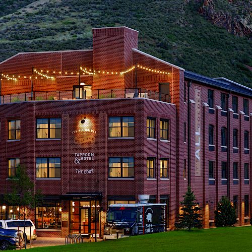 A brick building with string lights on the rooftop terrace. It is identified as a bar and hotel, surrounded by greenery and parked cars.