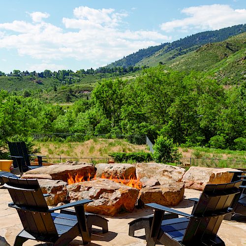 An outdoor seating area with a fire pit surrounded by nature, including trees and hills under a partly cloudy sky.
