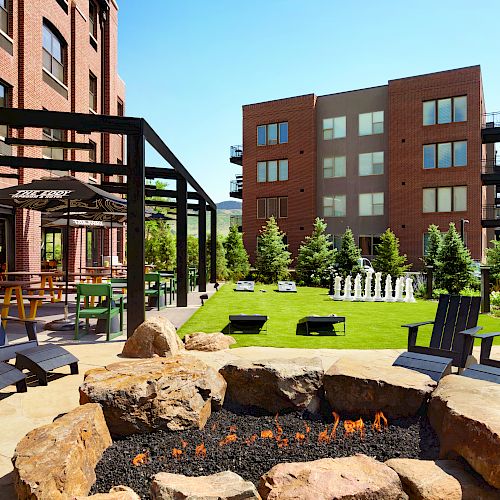 Outdoor area with a fire pit, chairs, a grassy lawn, trees, and a large chess set, located between modern brick buildings under a clear sky.